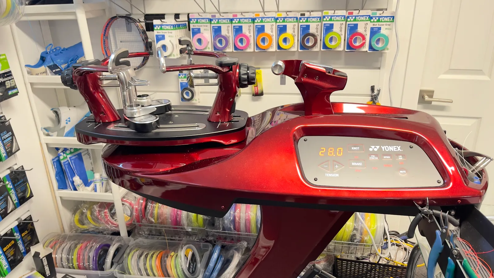A deep red Yonex electronic stringing machine, its panel reading 28.0, with the racket clamps and turntable to the left and reels of string on the shelf below.